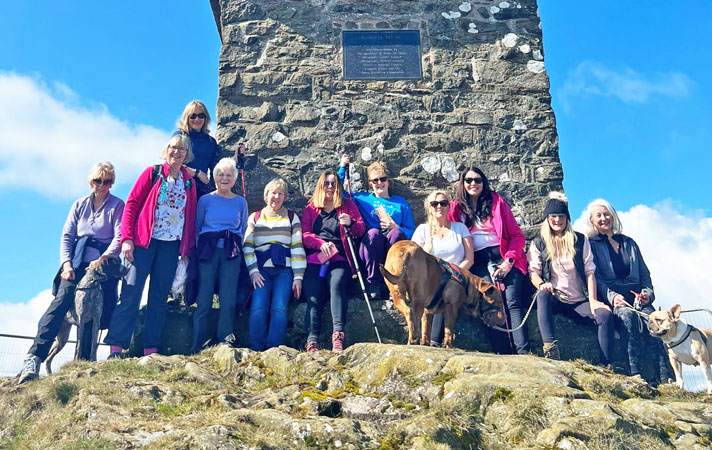 Eleven ladies with three dogs stand at the bottom of a stone pillar with blue skies behind