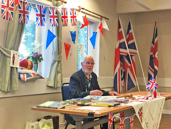 A man sits at a table ready to talk to the meeting with jubilee flags all around him
